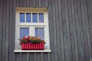A portion of gray house is pictured. It has a window with 2 windows that open and 4 grids on top. There is a red flower box hanging from window.